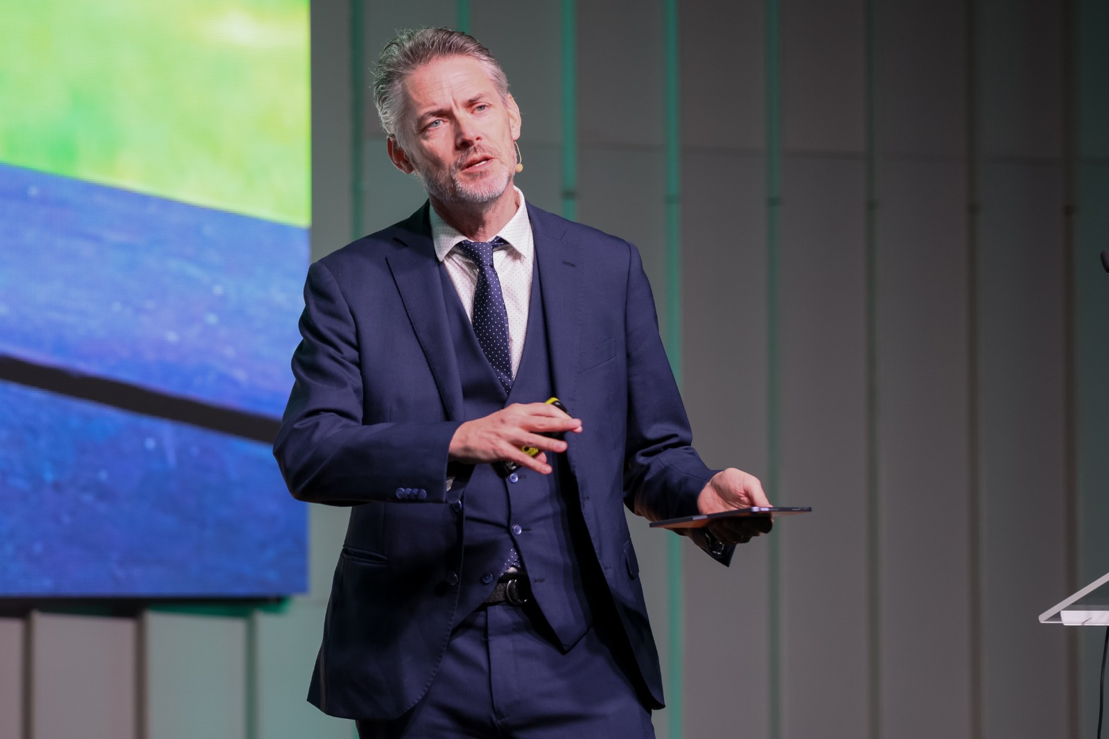James Cridland sur la grande scène de Radiodays Europe 2026 à Riga. Portrait en pied, costume trois-pièces bleu marine, durant sa keynote sur l'avenir de la radio et la connexion humaine.